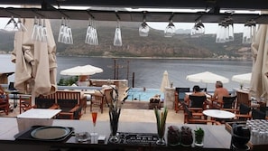 An outdoor seating area with tables and chairs overlooking a body of water, with umbrellas and a bar counter visible.