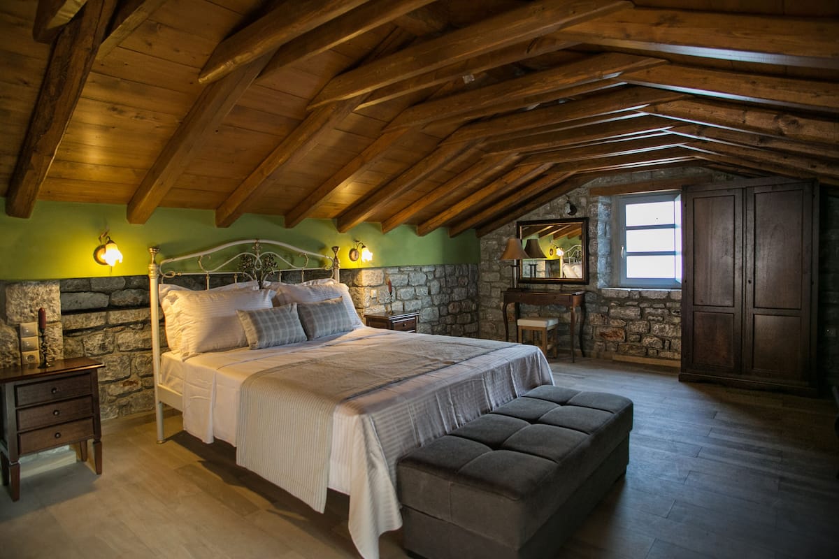 A bedroom with a stone wall, wooden ceiling beams, a bed with pillows, a bench, and a small table with a lamp.
