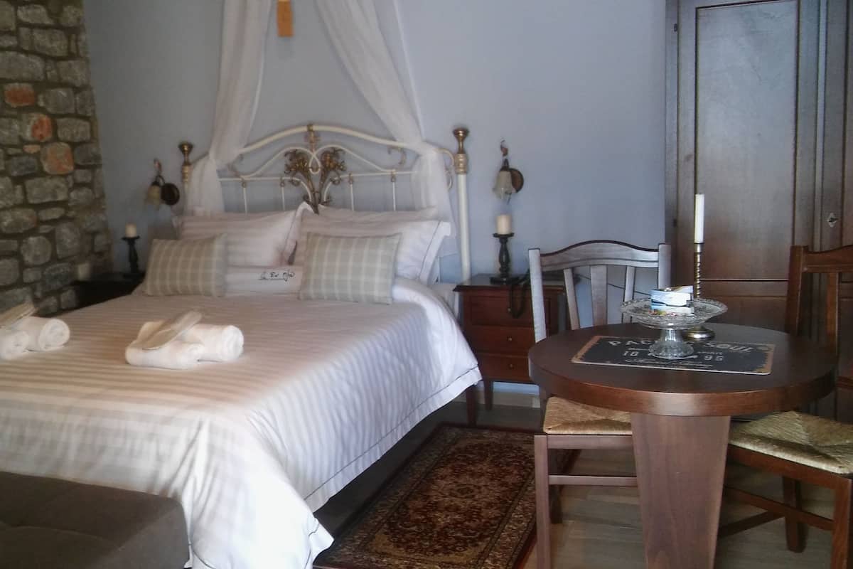 A neatly made bed with white linens and two towels, a wooden nightstand with a lamp, a wooden chair, and a small table with a coaster.