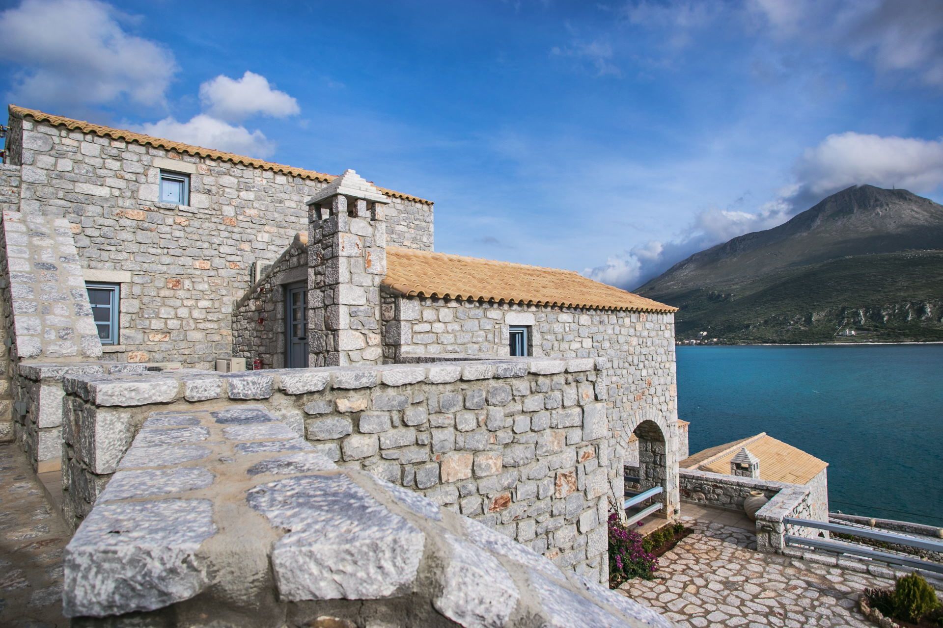 A stone building with a tiled roof overlooking a body of water and a mountain.