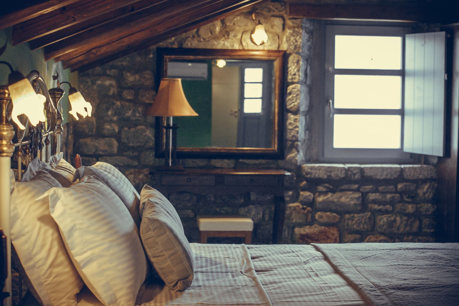 A cozy room with a stone wall, a bed with pillows, a lamp, and a window with shutters.