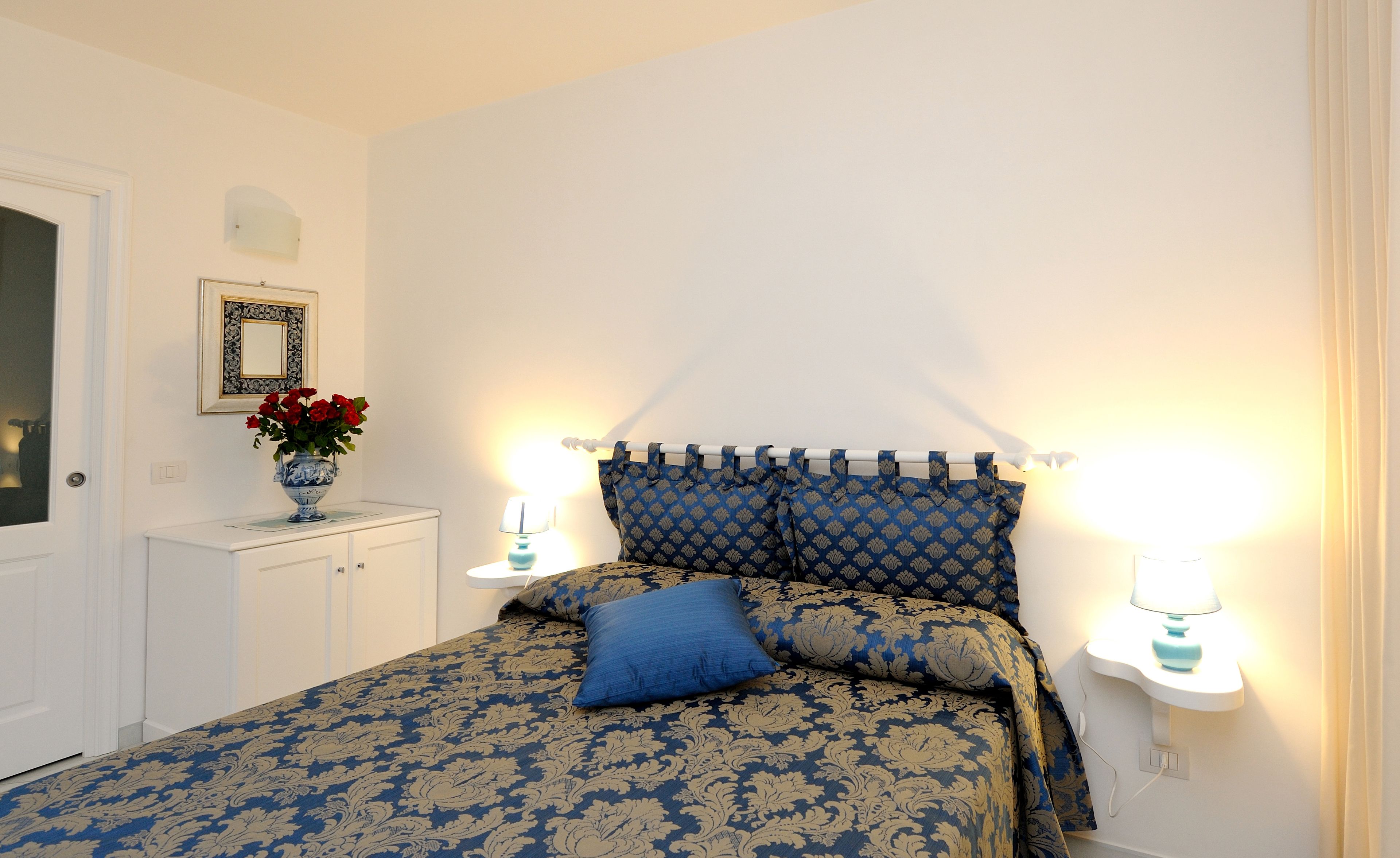 A bedroom with a bed, bedside lamps, a nightstand, a dresser, and a vase with flowers.