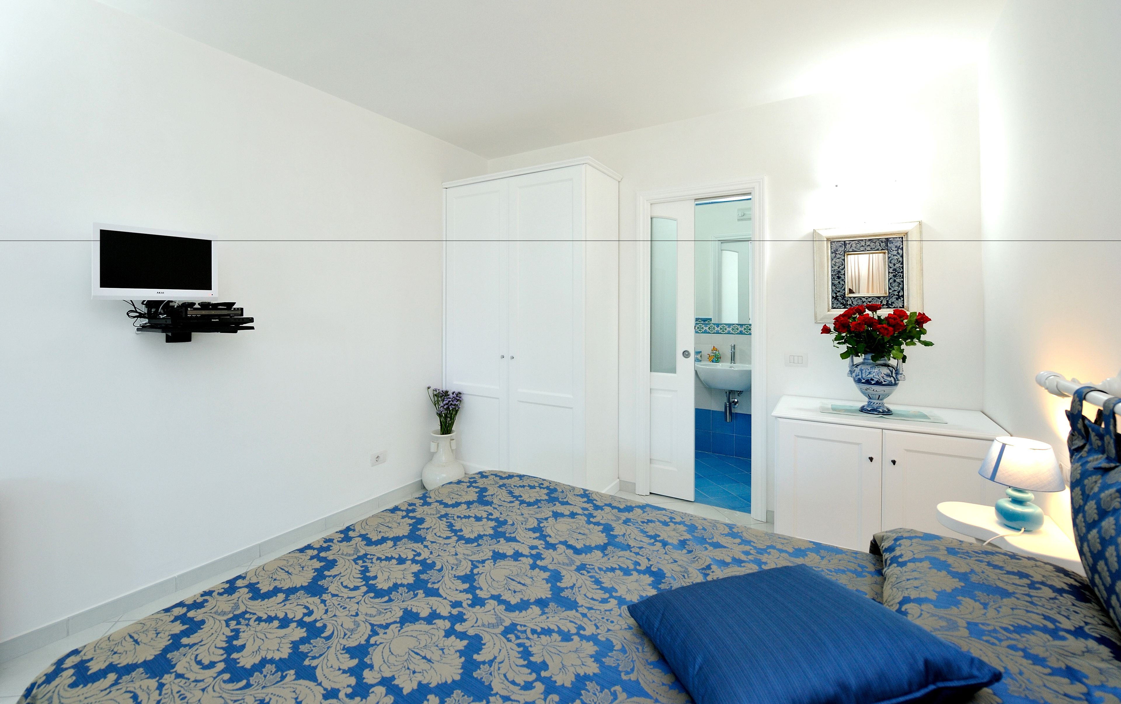 Un dormitorio con una cama, un televisor montado en la pared, una mesita de noche con lámpara y un baño visible a través de una puerta abierta.