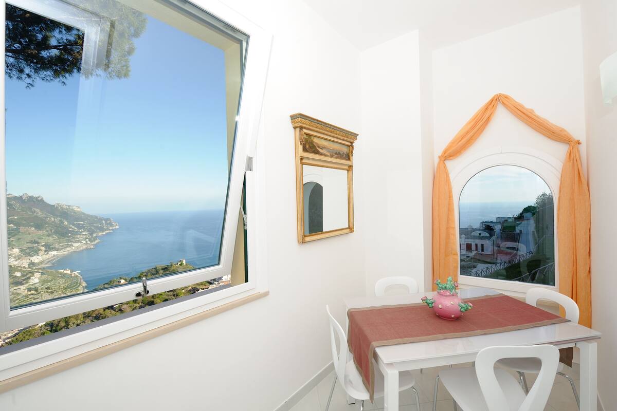 A room with a view of the sea, a table with chairs, a mirror, and a window with curtains.