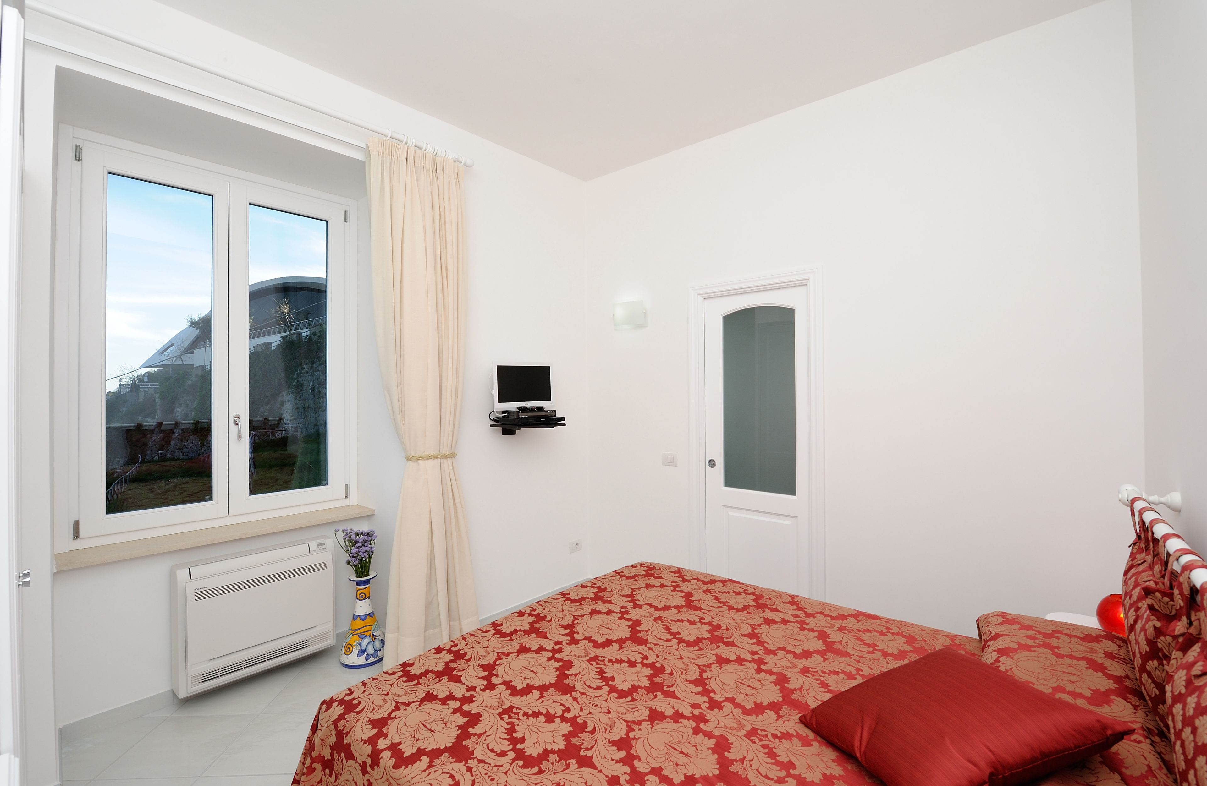 Een slaapkamer met een bed, een raam met gordijnen, een radiator, een tv aan de muur en een deur.