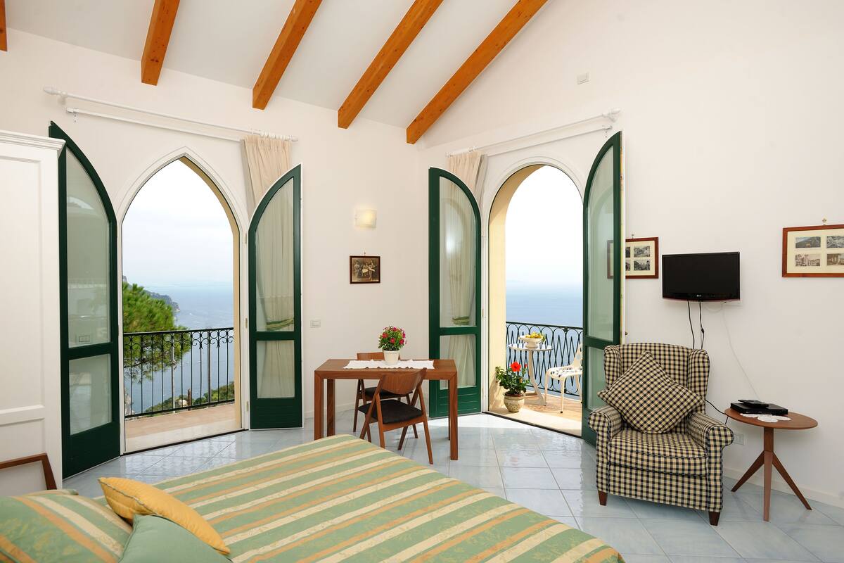 A bedroom with a bed, a chair, a table, and a balcony with a view.