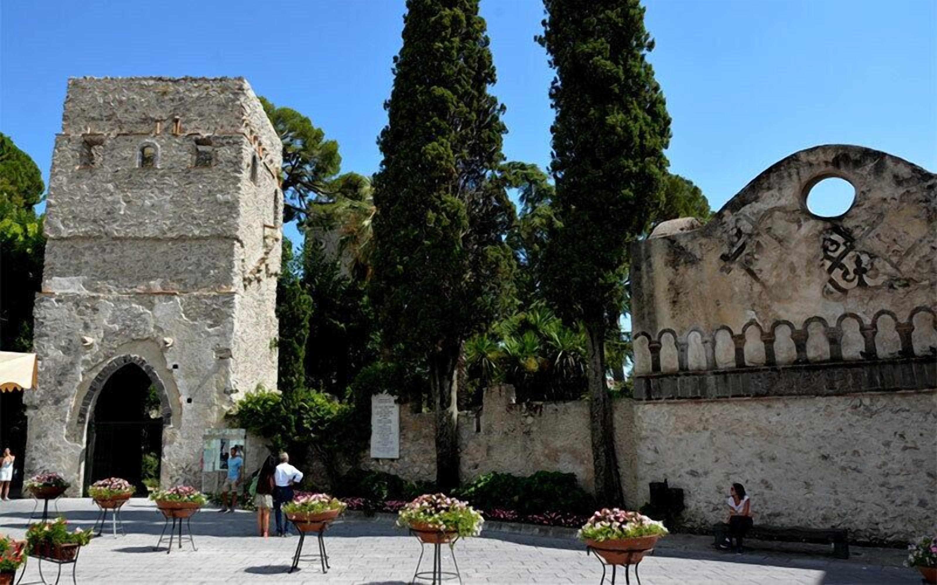 Um edifício histórico de pedra com uma torre alta e uma entrada em arco. Há vasos de flores em primeiro plano e uma grande parede com detalhes decorativos.