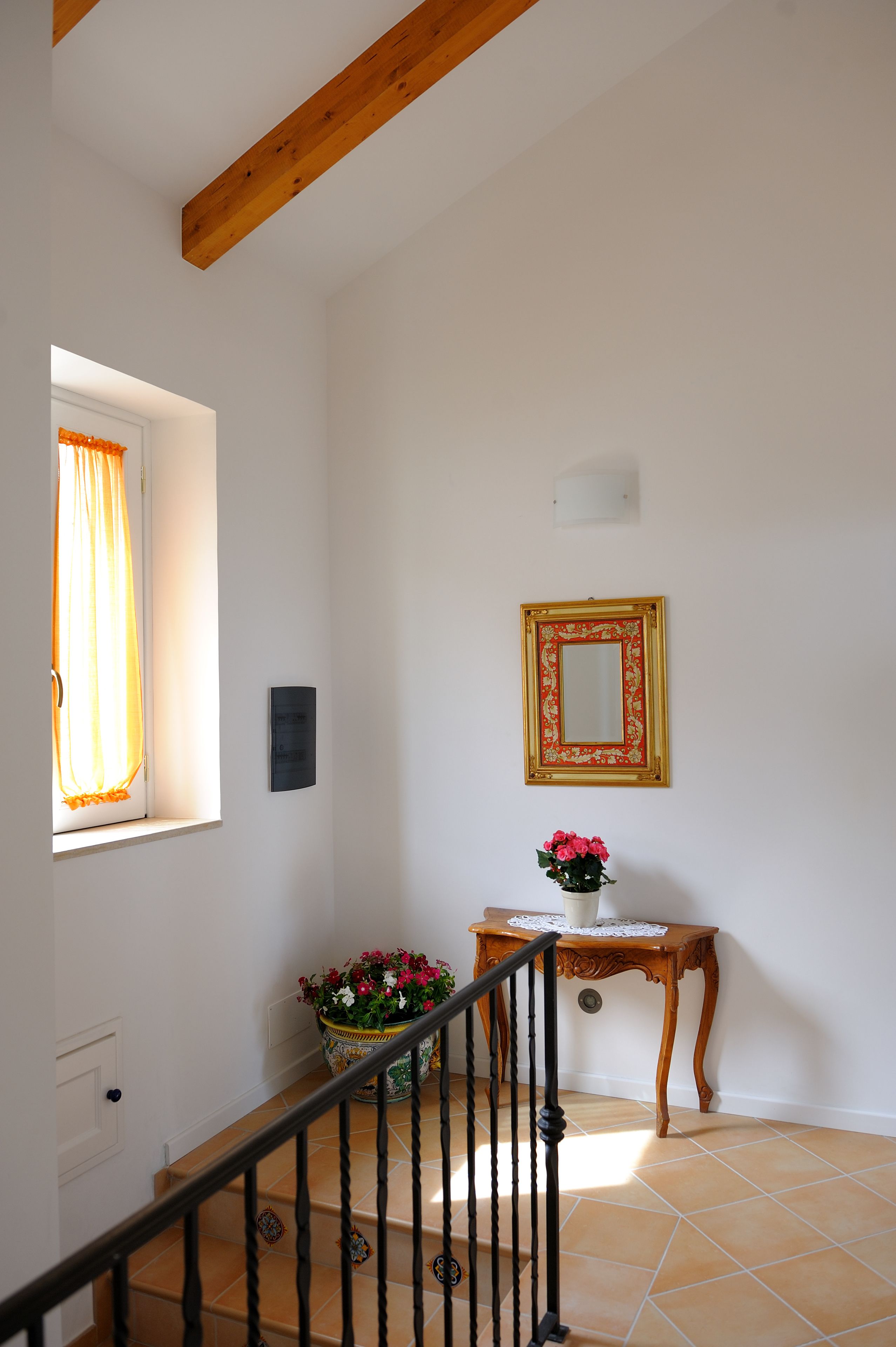 Una escalera con techo de vigas de madera, un espejo en la pared, una pequeña mesa con un florero y una ventana con cortinas.