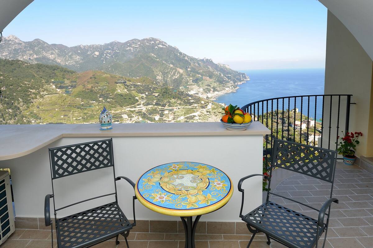 A balcony with a table and chairs, overlooking a coastal landscape with mountains and the sea.