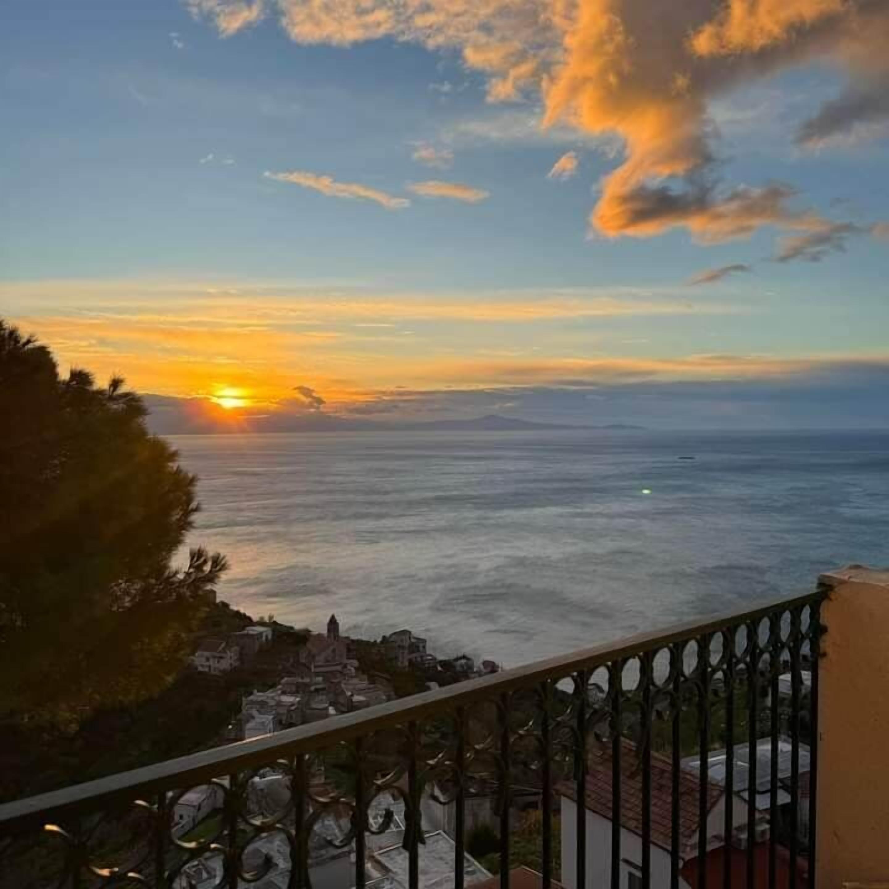 Kystudsigt med solnedgang, en balkon med gelænder og bygninger i det fjerne.