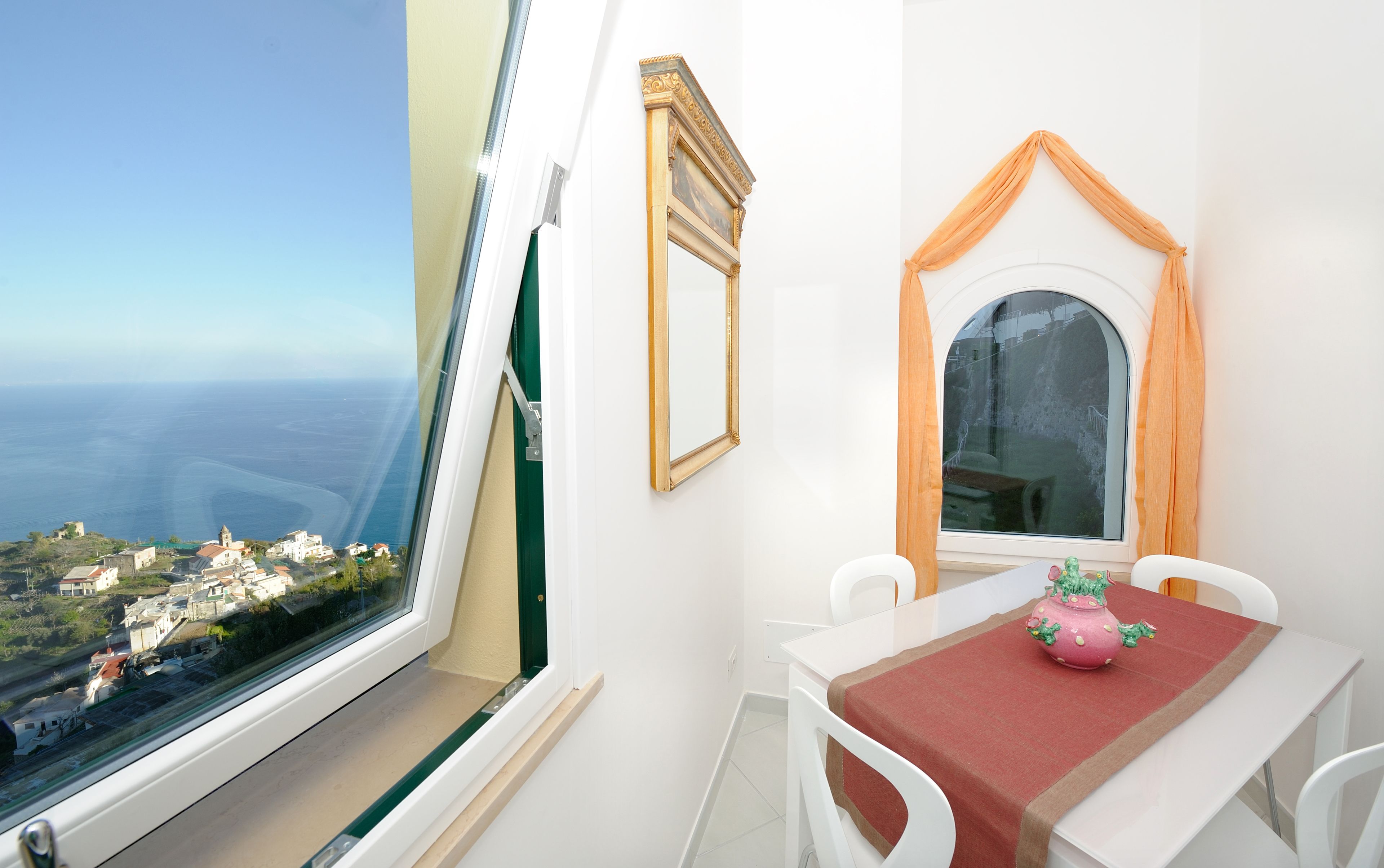 Ein Zimmer mit großem Fenster mit Blick auf eine Küstenstadt, einem kleinen Tisch mit einem rosa Topf und einem Spiegel mit aufwendigem Rahmen.