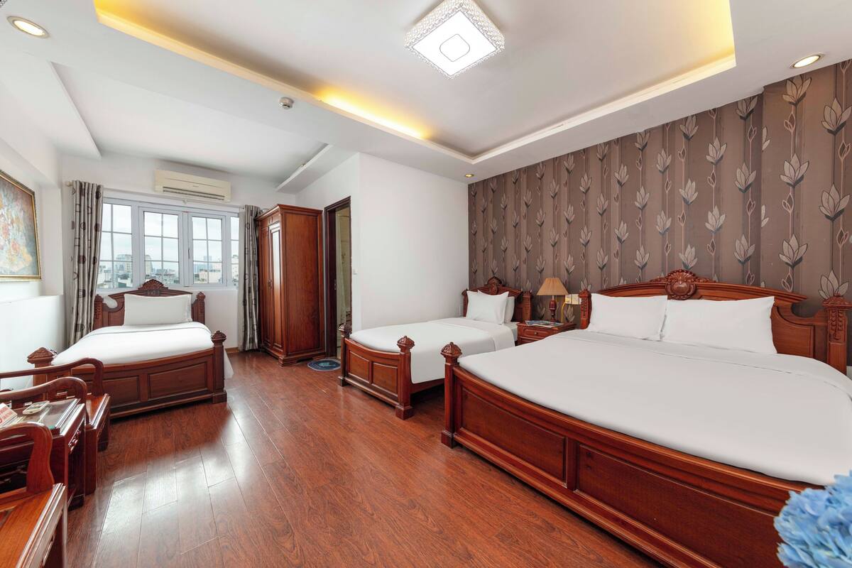 A hotel room with a large bed, two smaller beds, wooden furniture, a painting, and a view through the window.