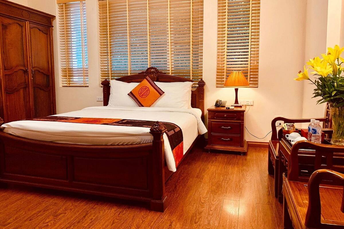 A traditional wooden bed with a headboard, a bedside table with a lamp, and a nightstand with a vase of flowers.
