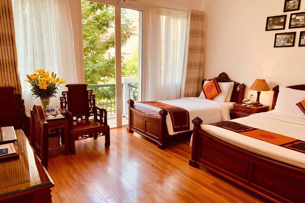 A hotel room with a wooden floor, two beds, a desk with chairs, a vase of flowers, and a balcony with a view of greenery.