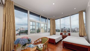 A hotel room with two beds, a wooden table with a vase of flowers, and a view of the city through large windows.