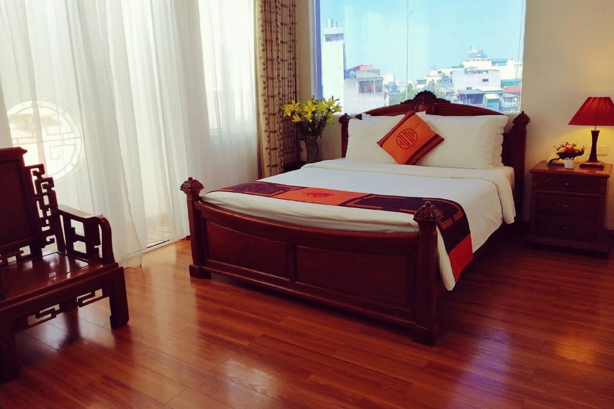 A hotel room with a wooden bed, a nightstand with a lamp, a chair, and a view of the city through large windows.