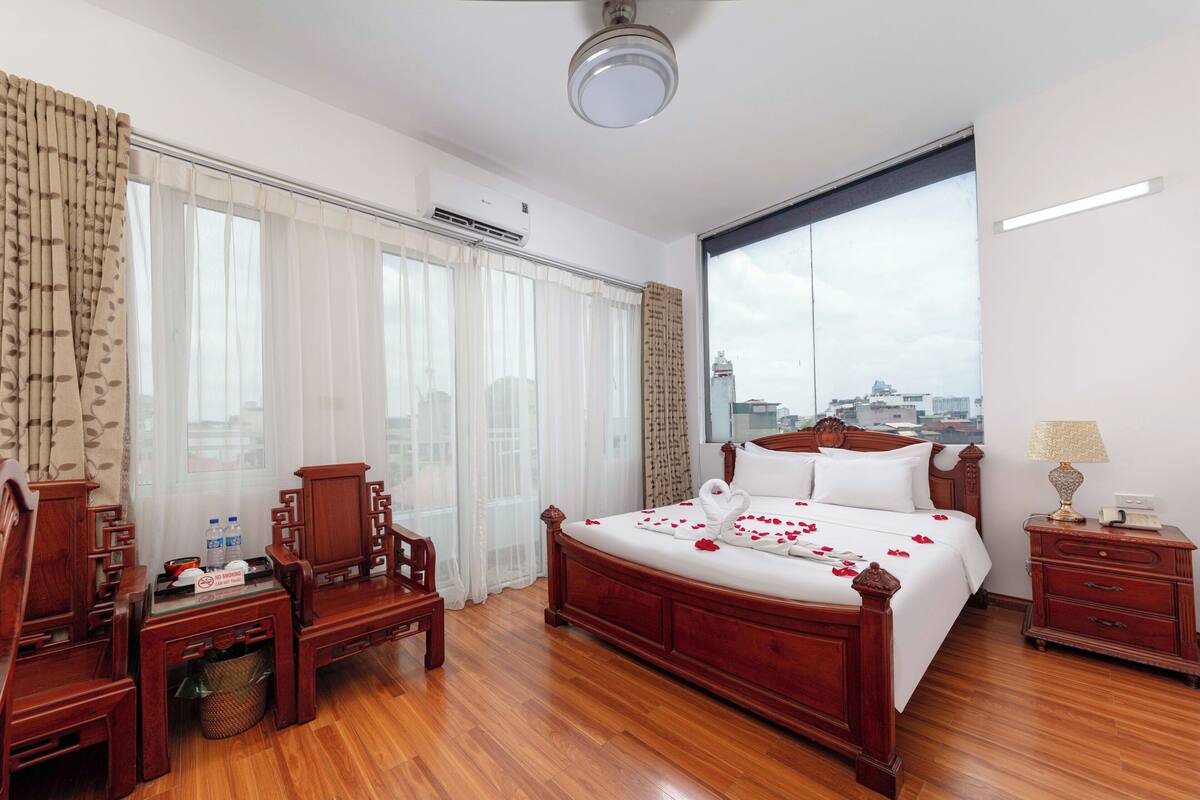 A hotel room with a large bed, wooden furniture, a desk, and a view of the city through a large window.