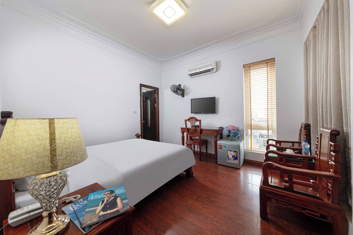 A hotel room with a bed, a desk, a chair, a TV, and an air conditioning unit.