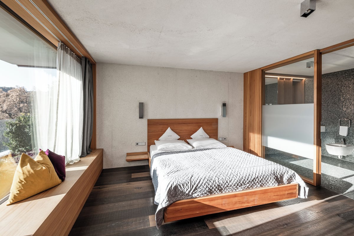 A modern bedroom with a wooden bed, a large window with curtains, a built-in bench, and a view of trees.