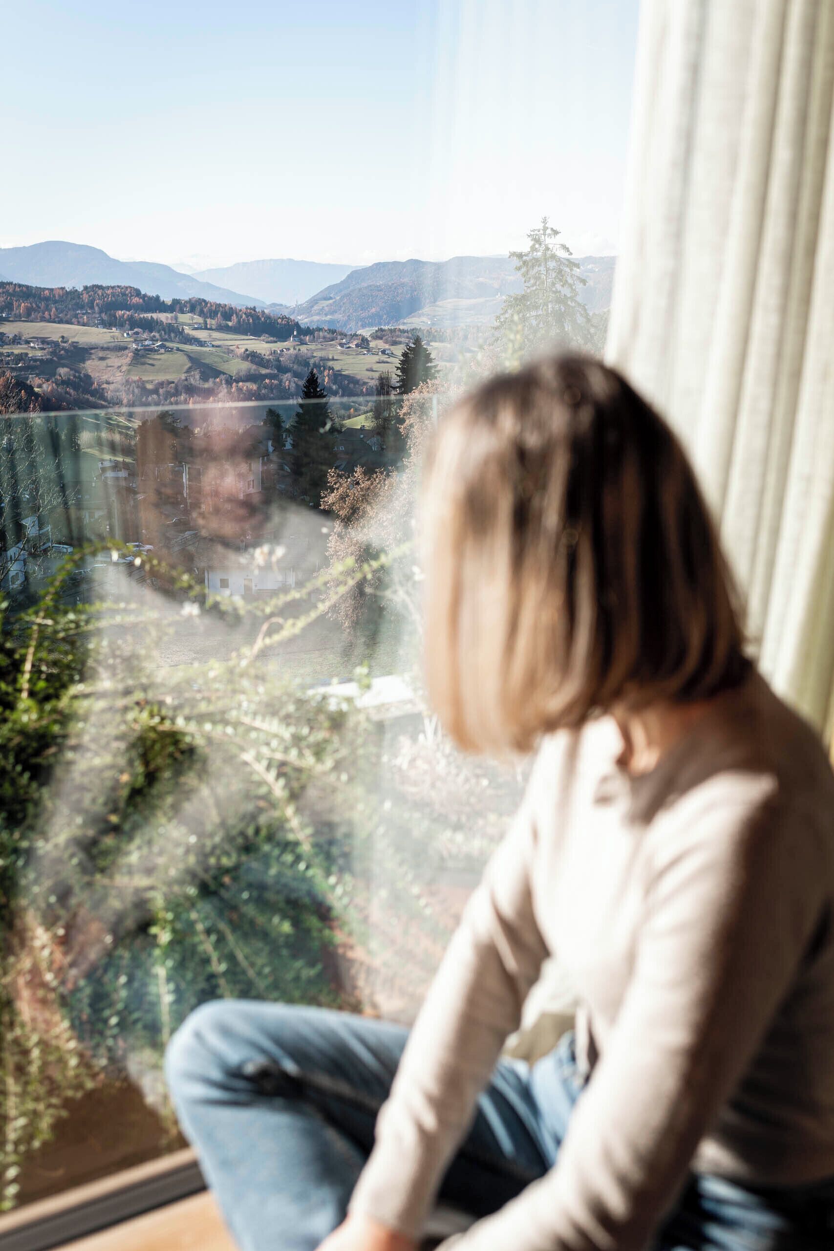 Una persona sentada junto a una ventana con vistas a un valle y montañas a lo lejos.