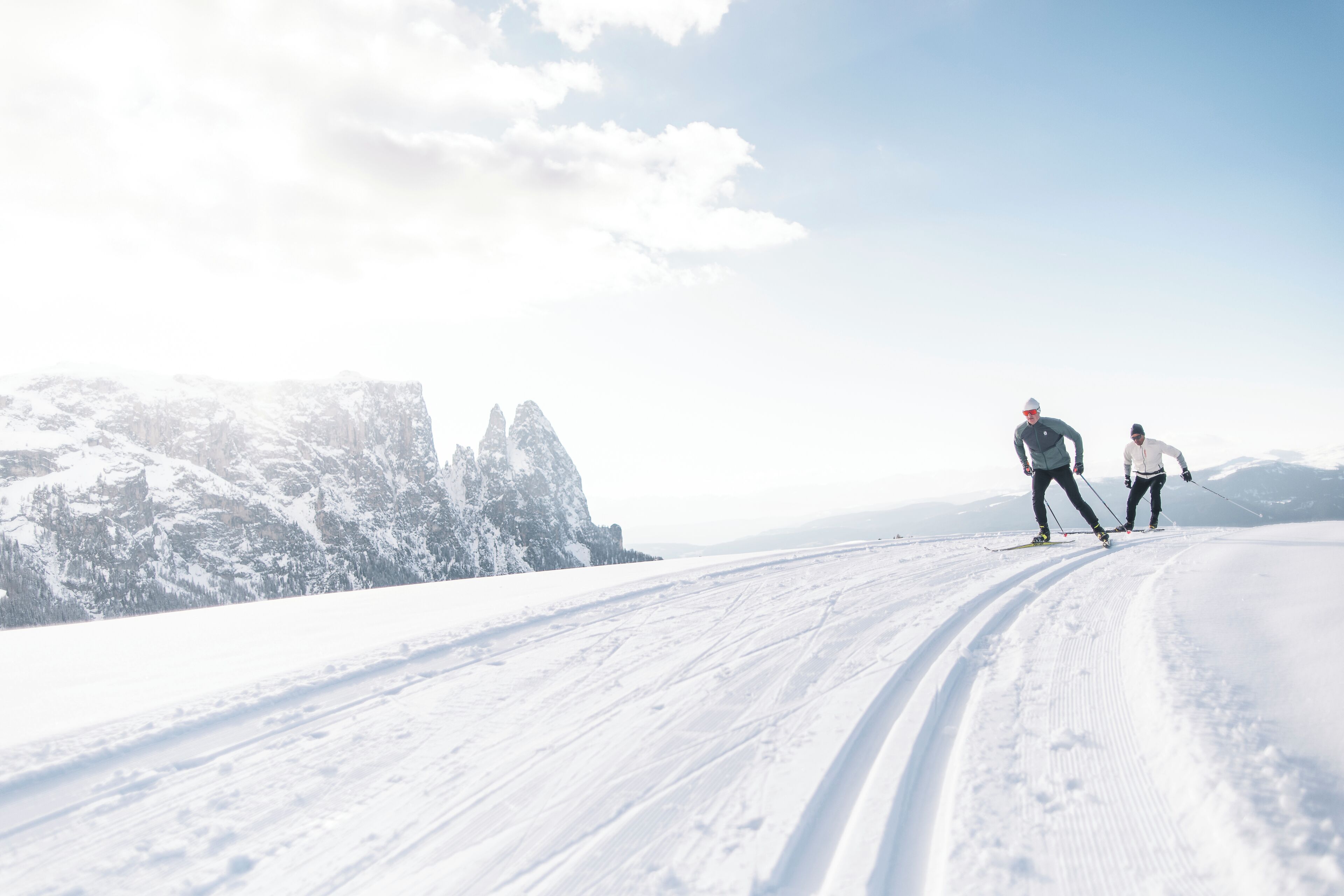 A snowy landscape with two skiers, a clear sky, and mountains in the background.