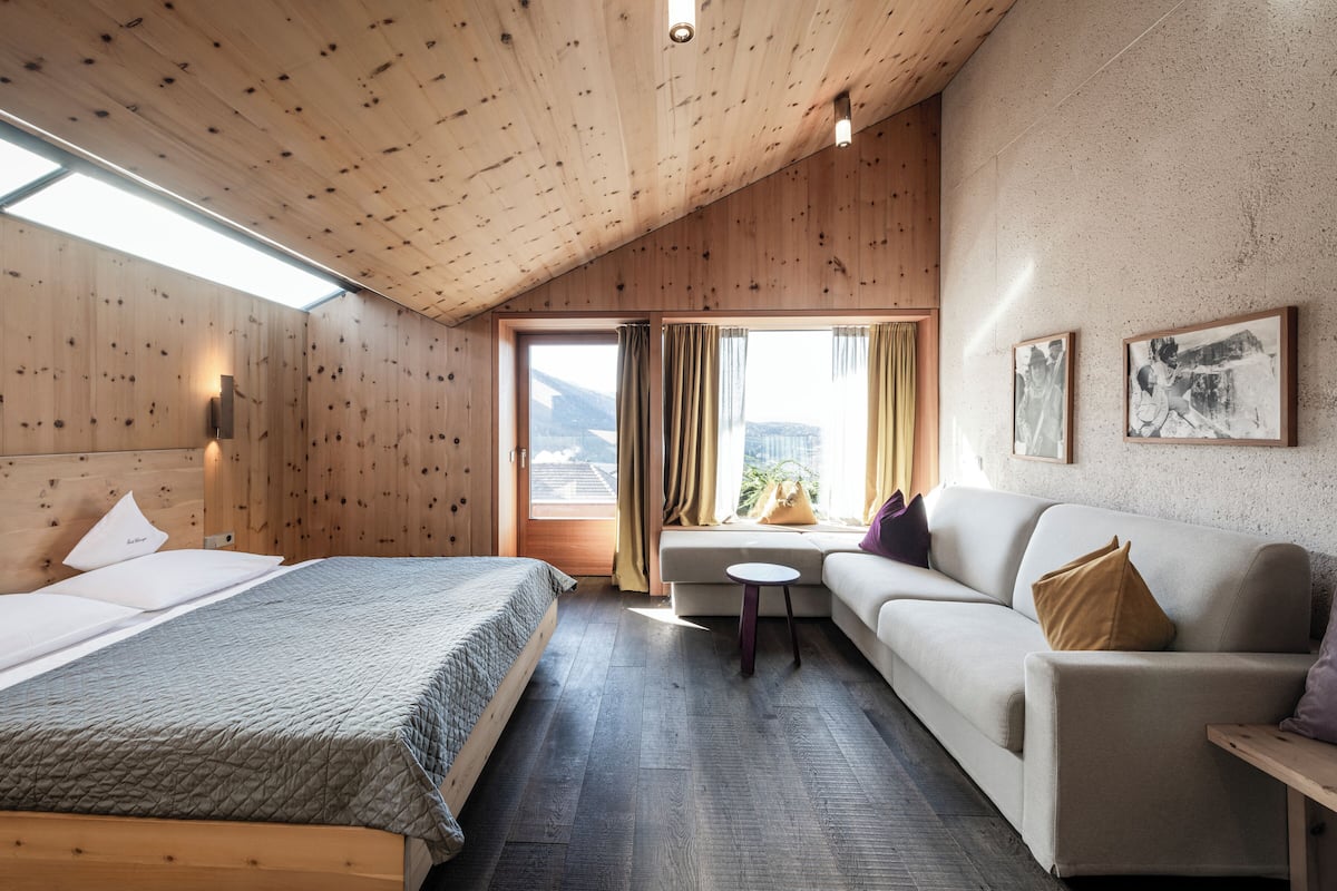 A room with a wooden ceiling, a bed, a sofa, a small table, and a window with curtains.