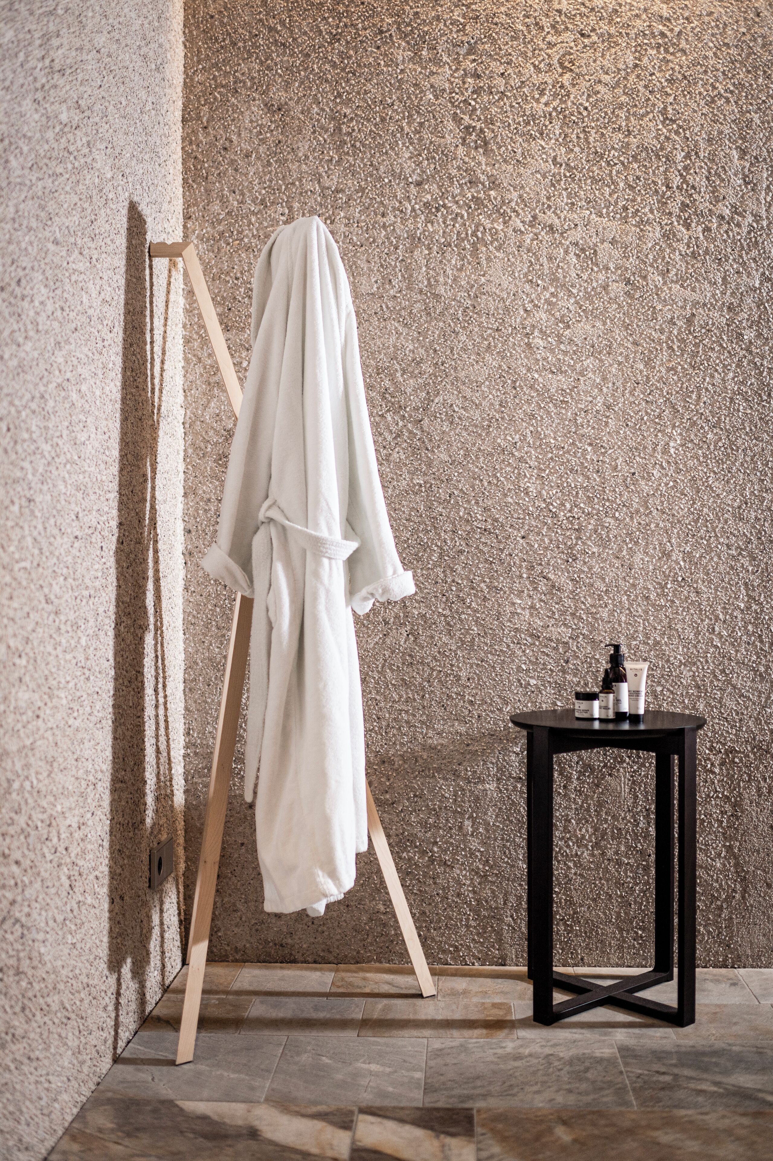 A bathroom with a textured wall, a wooden easel with a white towel, and a small black table with toiletries.