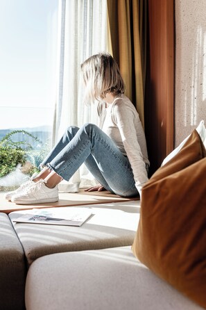 A person sitting on a sofa near a window with curtains, a magazine on the sofa, and a view of greenery outside.
