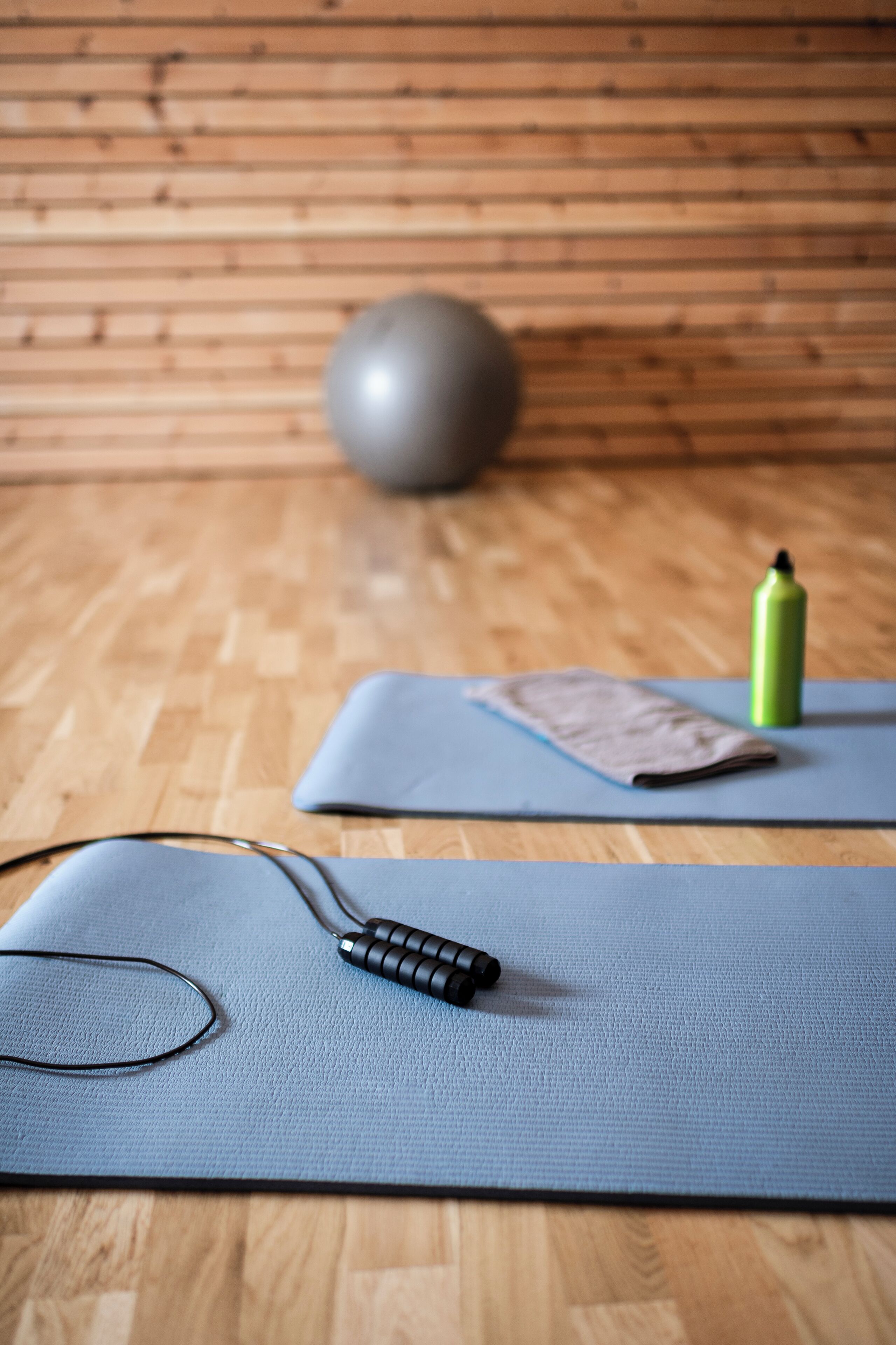 Ett trägolv med en blå träningsmatta, en yogamatta, ett par glasögon, ett svart föremål och en grön flaska.