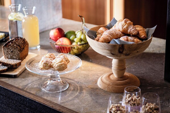 Desayuno con cruasanes, muffins y un bol de fruta sobre una encimera de mármol.