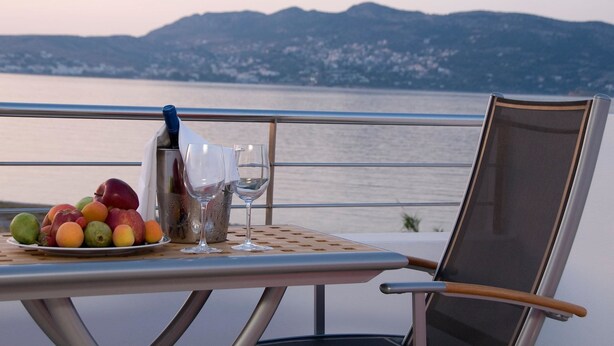 A table with a fruit plate, a bottle, and glasses, overlooking a body of water with mountains in the background. - Sea Sight Hotel (Markopoulo Mesogaias)