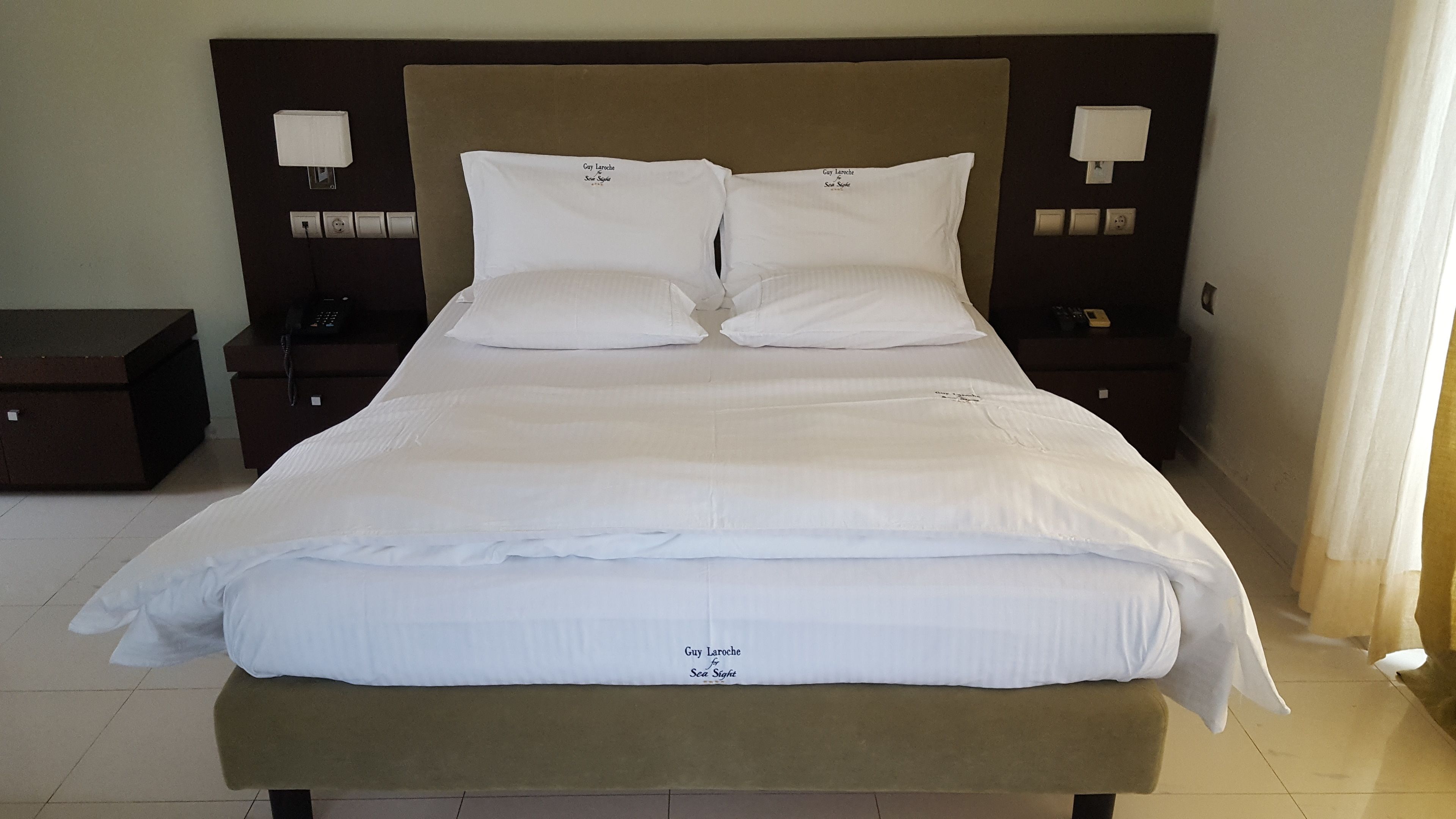 Un lit bien fait, avec du linge de lit blanc et des oreillers, une tête de lit marron et des lampes de chevet.