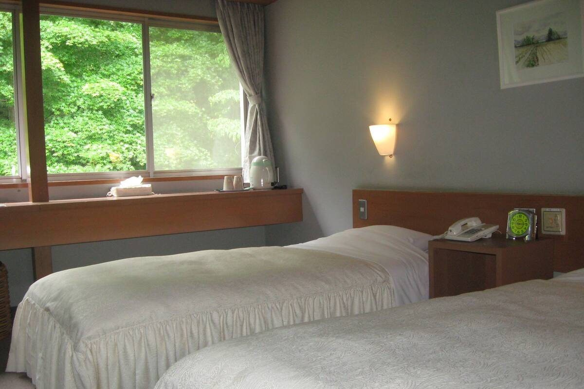 Quarto de hotel com duas camas, janela com cortinas, mesa de cabeceira com abajur, relógio e quadro pendurado na parede.