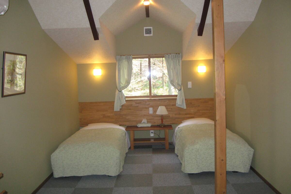 Quarto com duas camas de solteiro, janela com cortinas, escrivaninha com abajur e luminária no teto.