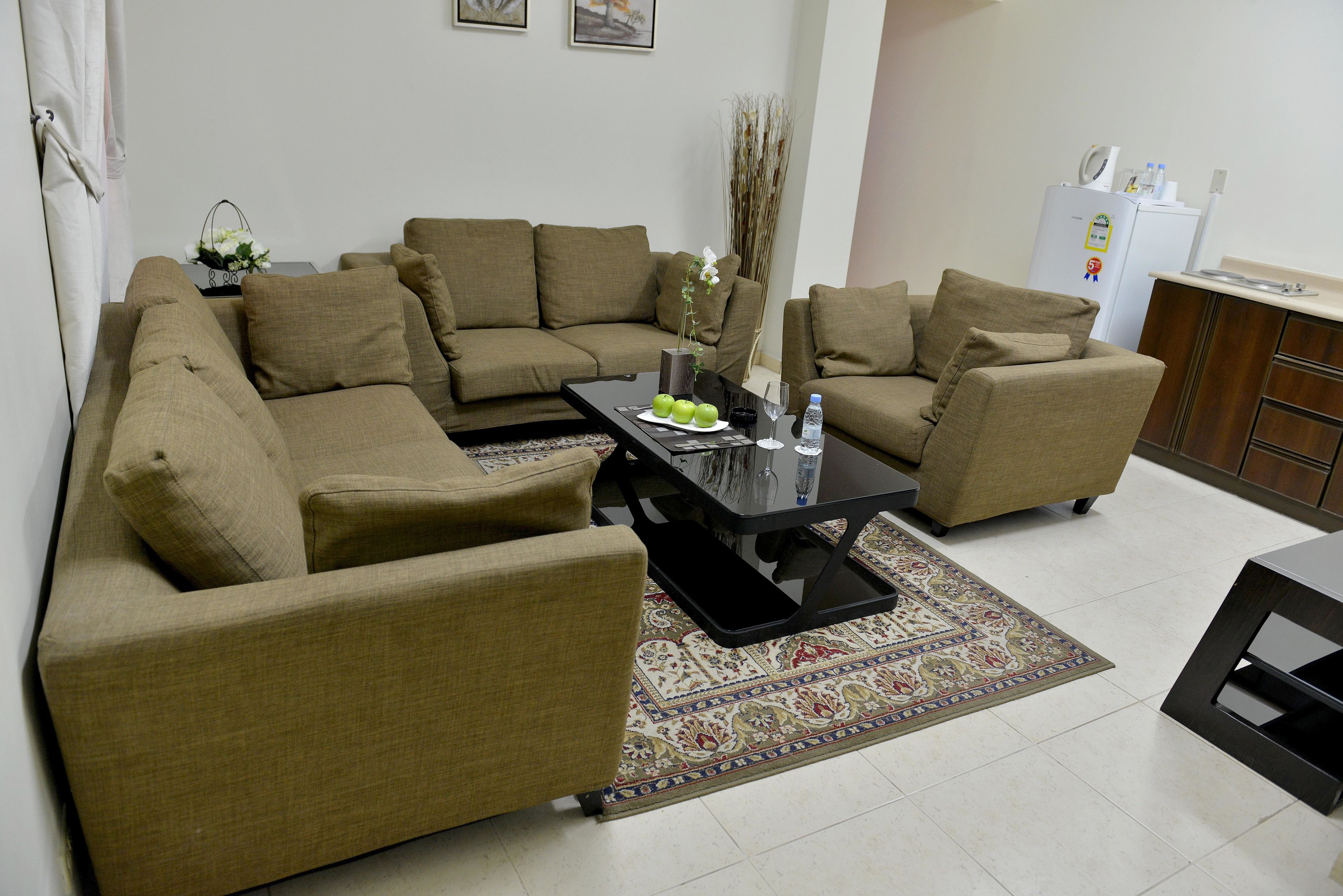 A living room with a sectional sofa, a coffee table, and a small kitchen area with a refrigerator.