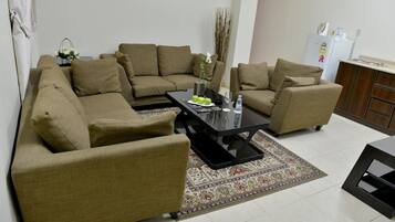 A living room with a sectional sofa, a coffee table, and a small kitchen area with a refrigerator.