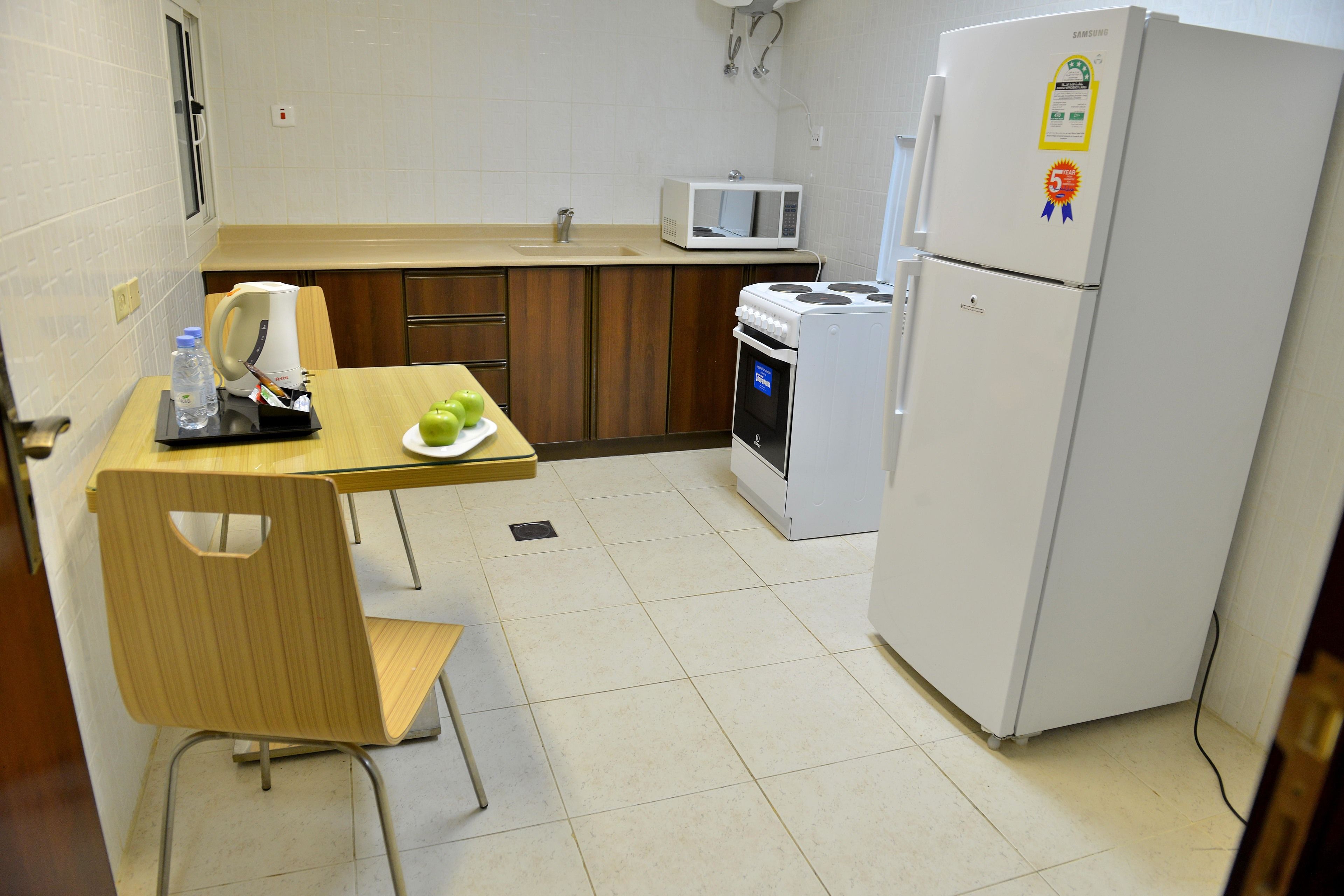 A small kitchenette with a microwave, stove, and refrigerator. There is a wooden table with a chair, a kettle, and a plate with green apples.