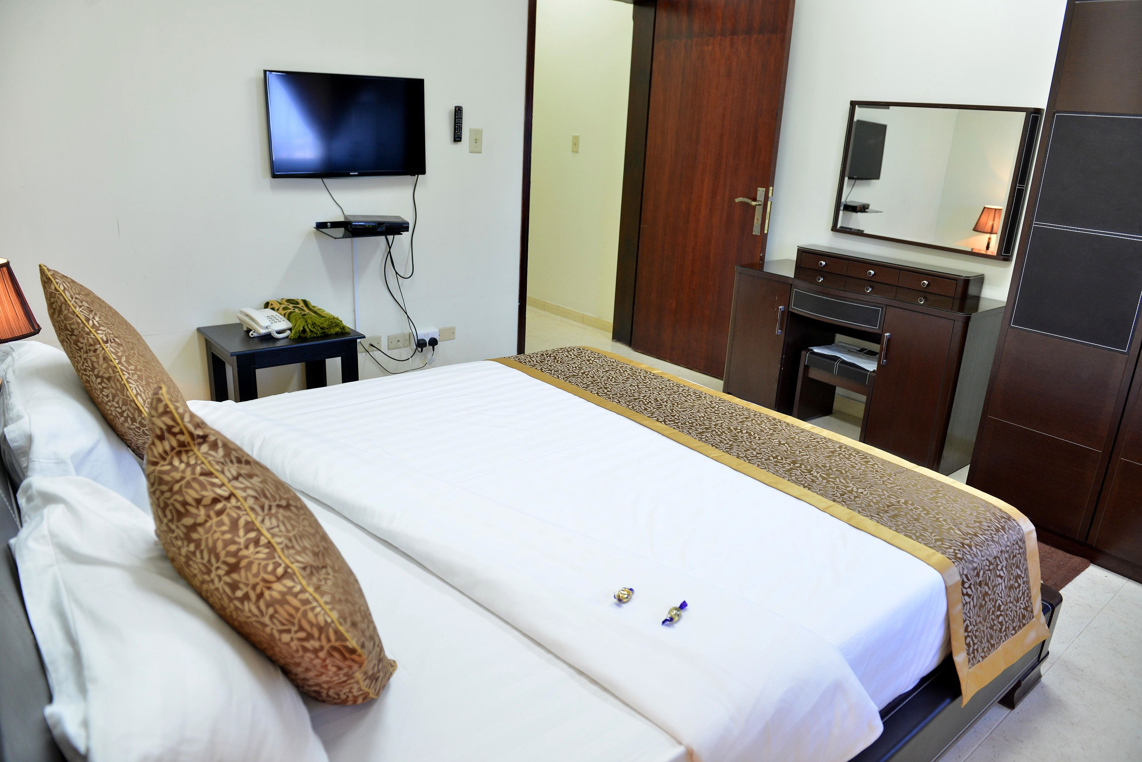 Habitación de hotel con una cama, televisor montado en la pared, una mesita con un teléfono y una planta, y un armario con espejo.