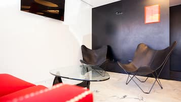 Ein modernes Interieur mit einem Glastisch, zwei Ledersesseln, einem roten Sofa und einer dunklen Wand mit einer Leuchte.