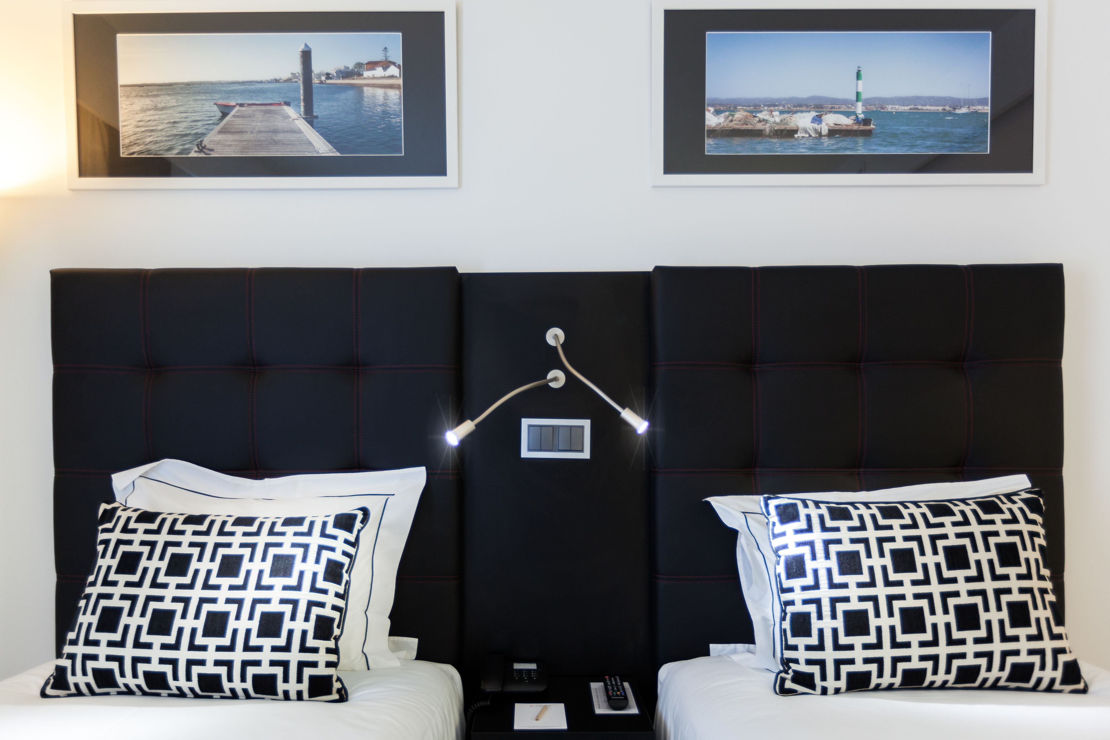 A hotel room with a dark headboard, two beds with matching pillows, a bedside table with a lamp, and two framed pictures on the wall.