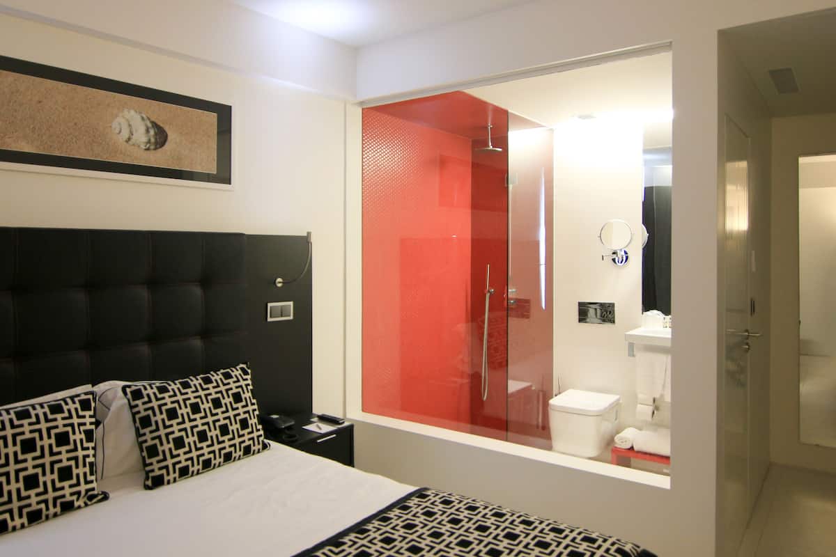 A hotel room with a bed, a nightstand, a bedside table, a red shower enclosure, a bathroom visible through the shower door, and a framed picture on the wall.