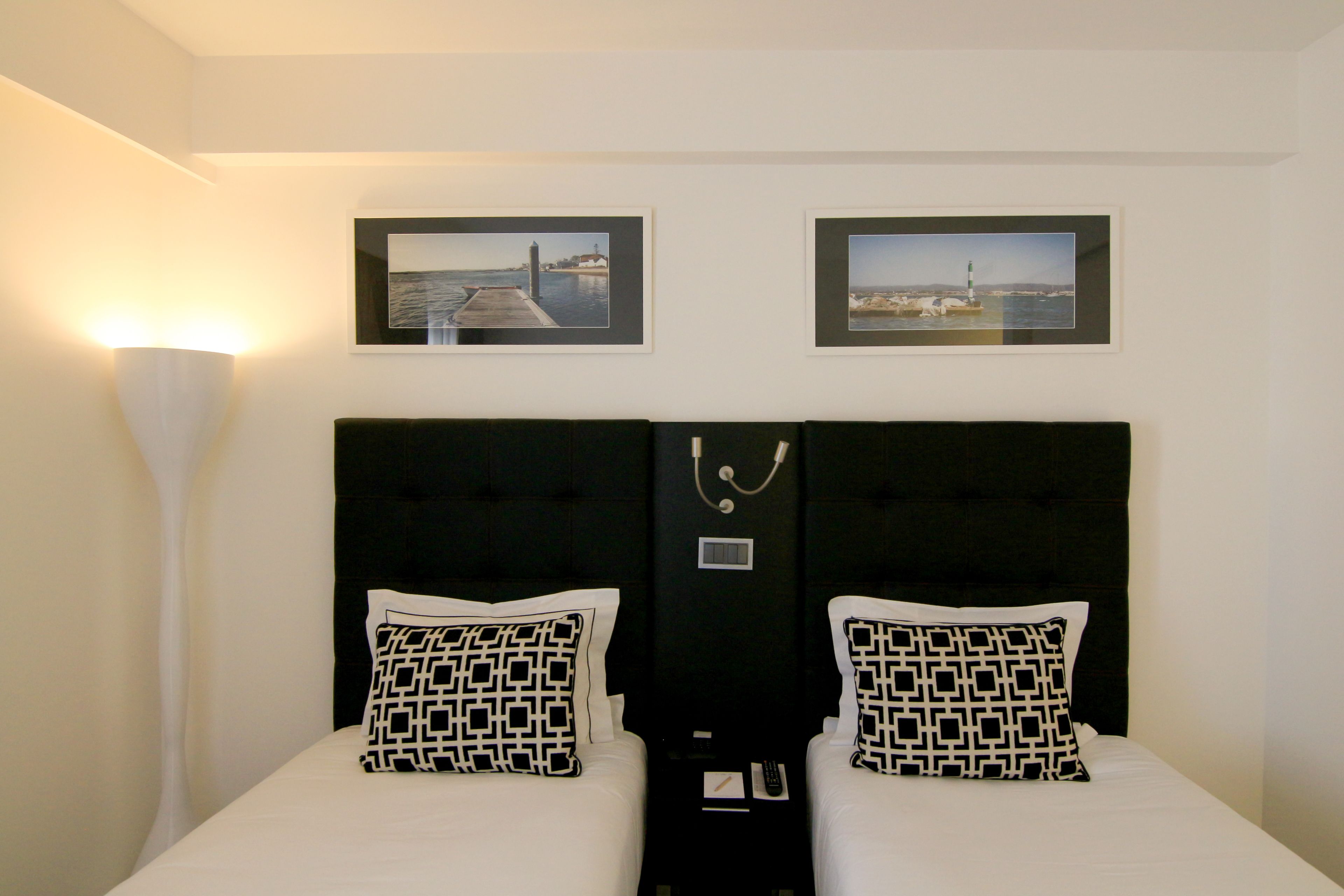 Quarto de hotel com duas camas, cabeceira com painel de controle e duas imagens emolduradas na parede.