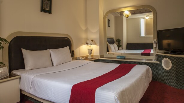 A hotel room with a bed, a TV, a mirror, a nightstand, and a lamp. - Good Hotel (Taichung)