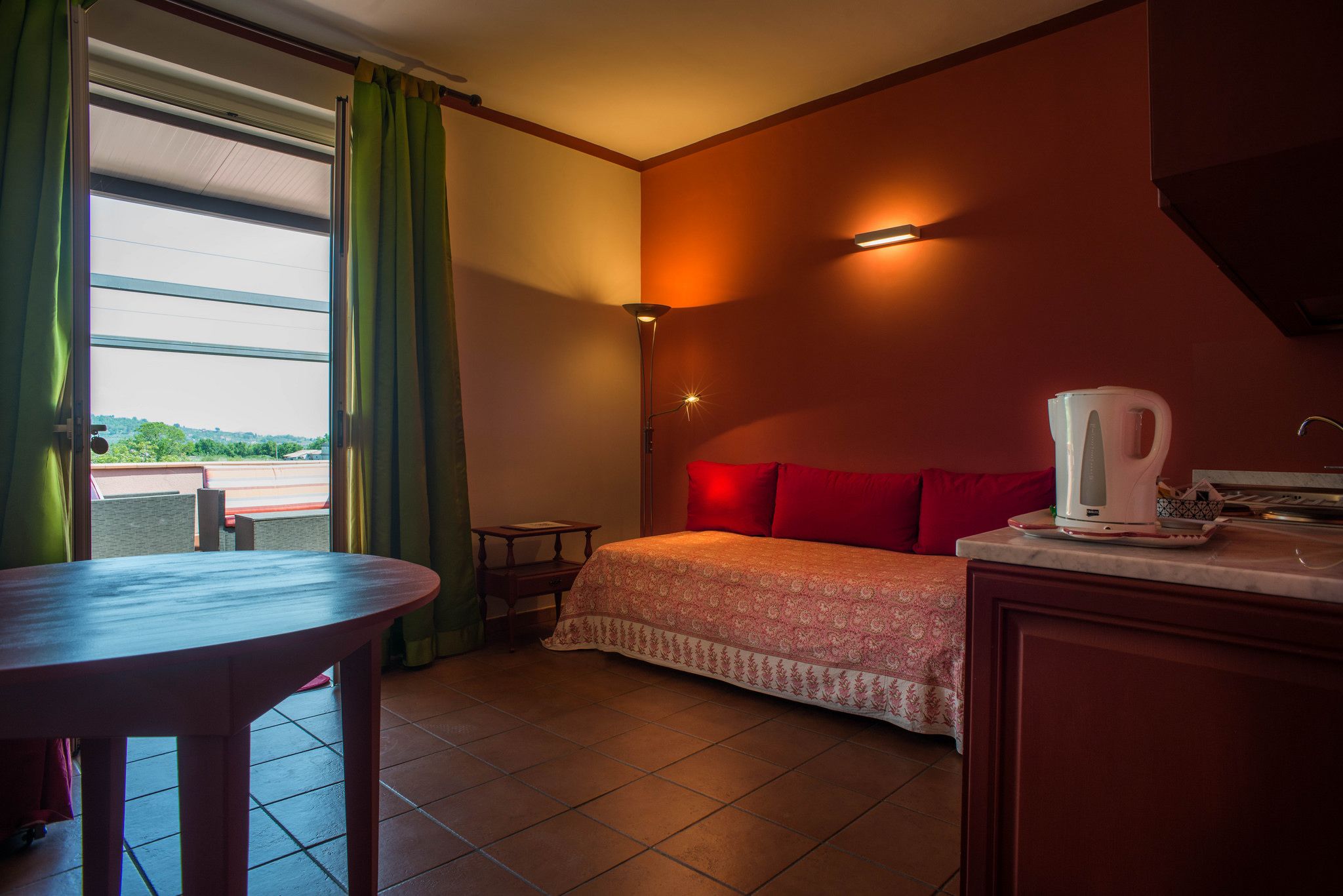 A hotel room with a bed, a table, a kettle, and a view of the outdoors.