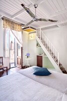 Un dormitorio con una cama, una silla, una mesita con un florero, una escalera y un ventilador de techo.
