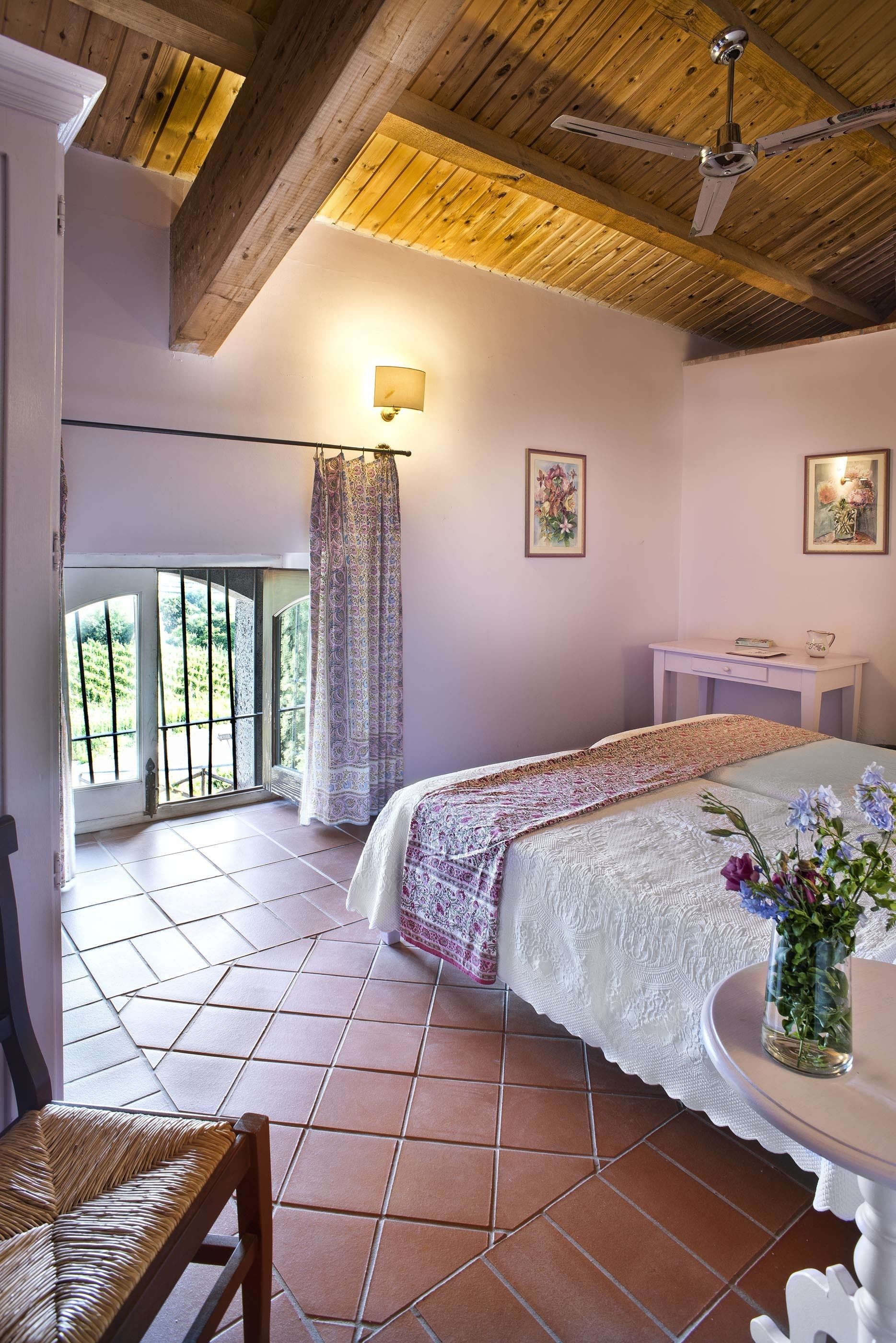 A bedroom with a bed, a desk, a chair, a ceiling fan, a window with a view, and a wall-mounted lamp.