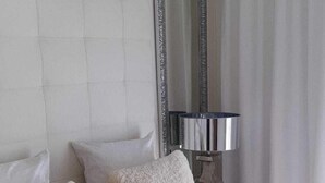 Een net opgemaakt bed met hoofdeinde, twee kussens, een bedlampje en een spiegel op een nachtkastje.