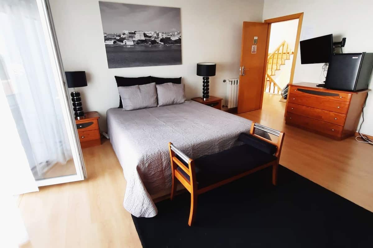 A bedroom with a bed, bedside tables, a dresser, a television, and a cityscape painting on the wall.