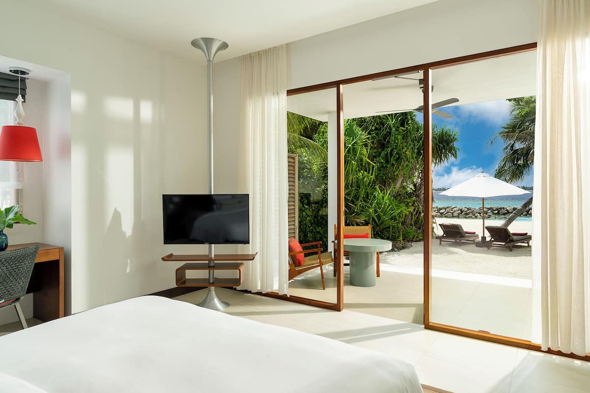 A hotel room with a bed, a TV, a desk, and a view of a beach with palm trees and a white umbrella.