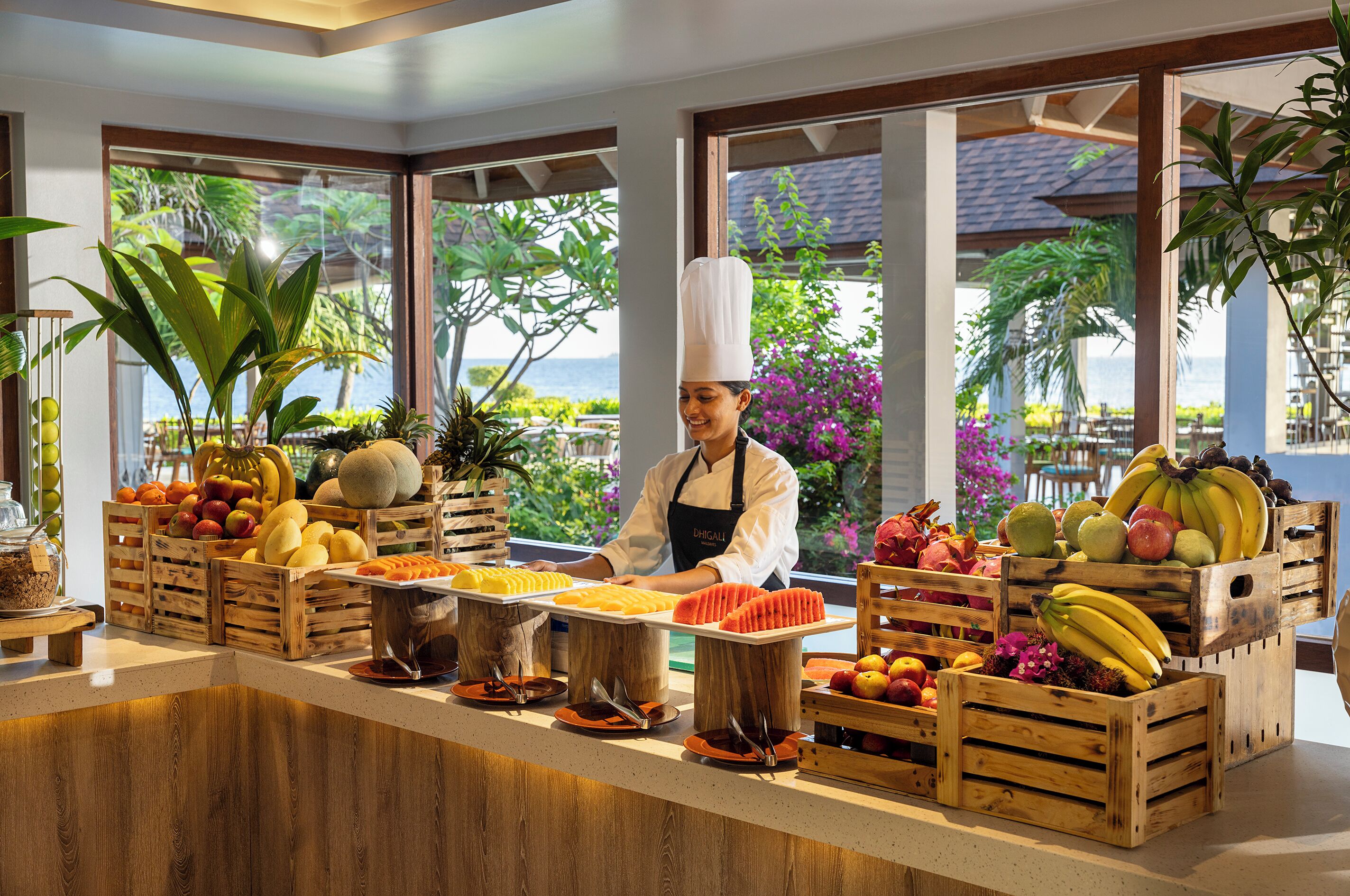 A buffet counter with various fruits and vegetables, a chef in uniform, and a view of the outdoors through large windows.